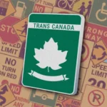 Зеленый прямоугольный знак с белой надписью "TRANS CANADA" вверху и белым кленовым листом на зеленом фоне с декоративной лентой. Знак "Trans-Canada Highway" обозначает маршрут Трансканадского шоссе — главной национальной автомагистрали Канады